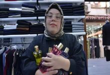 Fotoğrafta başörtülü ve gözlüklü genç bir kadın, bir giyim mağazasının içinde kollarında birkaç parfüm şişesi tutarak kameraya doğru duruyor. Arka planda raflara dizilmiş pantolonlar ve askıda kıyafetler görülüyor; mağaza düzenli ve aydınlık bir çarşı dükkânı atmosferi veriyor.