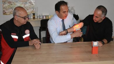 Bir masa etrafında üç erkek oturuyor. Ortadaki kişi, yanındaki adamın koluna tuttuğu cihazla damarları görünür hale getiriyor; cihazın yaydığı ışıkla kol üzerinde damarlar belirgin şekilde parlıyor. Soldaki kişi ise dikkatle süreci izliyor. Ortam, bir eğitim ya da tanıtım anını andırıyor.