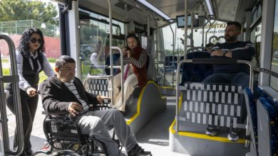 Kadın personel, tekerlekli sandalyedeki erkek yolcuyu otobüsün içine doğru yönlendirirken, içeride diğer özel gereksinimli yolcular koltuklarda oturuyor.