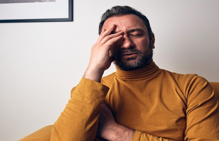 Bir koltukta oturan adam, gözlerini kapatmış şekilde alnını tutuyor. Yüz ifadesi yorgunluk, stres ya da duygusal bir sıkıntıya işaret ederken, sahne zihinsel yük ve içsel bir mücadele hissi veriyor.