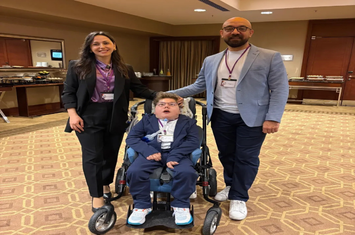 Fotoğrafta, bir otel lobisi veya etkinlik salonu olduğu anlaşılan mekanda, tekerlekli sandalyedeki bir çocukla birlikte duran iki yetişkin görülüyor.