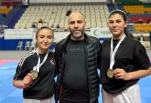 Bir spor salonunda çekilmiş fotoğrafta, ortada bir erkek ve iki yanında spor kıyafetli iki kadın yer alıyor. Kadın sporcular boyunlarında madalya taşıyor ve kameraya gülümseyerek poz veriyor. Arka planda tribünler ve minderli bir müsabaka alanı görülüyor.