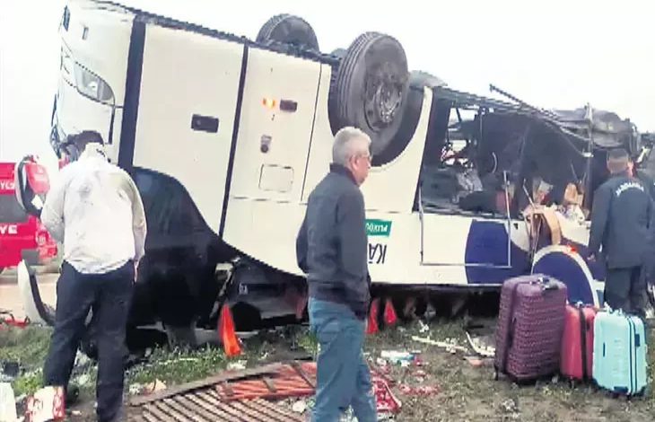 Görselde, devrilmiş bir yolcu otobüsü ve çevresinde inceleme yapan kişiler görülüyor. Otobüs yan yatmış durumda; bagajlar, kişisel eşyalar ve kırılmış parçalar etrafa dağılmış. Olay yeri görevlileri ile bazı siviller kazanın olduğu alanda inceleme yapıyor. Genel atmosfer acil durum müdahalesinin sürdüğünü gösteriyor.