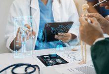 Bu görselde, bir sağlık çalışanı tablet kullanarak dijital bir sistem üzerinden insan iskeleti ve organlarını incelemekte, aynı zamanda masanın üzerinde küçük bir iskelet modeli ile insan anatomisi hakkında açıklama yapılmaktadır.