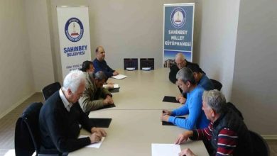 Bir toplantı salonunda çekilen fotoğrafta, uzun bir masa etrafında oturan birkaç yetişkin kişi Braille alfabesi (kabartma yazı) öğrenmek için önlerindeki kâğıt ve kabartma yazı tabletleri üzerinde çalışıyor. Arka tarafta Şahinbey Belediyesi ve Şahinbey Millet Kütüphanesi yazılı afişler yer alıyor. Katılımcılar sessiz bir ortamda not alarak eğitime odaklanmış görünüyor.