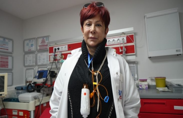 Görselde, bir sağlık kuruluşunun acil servis biriminde görevli bir sağlık çalışanı yer alıyor. Beyaz önlük giymiş olan kişi, üzerinde çeşitli tıbbi ekipmanlar taşıyor. Arka planda monitörler, tıbbi cihazlar ve acil müdahale malzemelerinin bulunduğu bir çalışma alanı görünüyor. Atmosfer, yoğun bir sağlık hizmeti ortamını yansıtıyor.