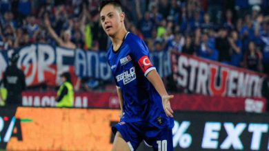 Bir futbol maçında, mavi formalı bir oyuncu sahada koşarken görülüyor. Kolunda kaptanlık bandı var ve üzerinde Türk bayrağı detayı dikkat çekiyor. Tribünler dolu, arka planda coşkulu taraftarlar ve pankartlar yer alıyor. Oyuncu gol ya da önemli bir pozisyon sonrası sevinç anı yaşıyor gibi görünüyor.