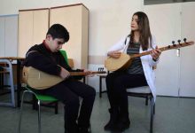 Bir sınıf ortamında, sandalyede oturan bir çocuk bağlama çalmaya çalışırken yanında oturan bir kadın da bağlama çalarak ona eşlik ediyor. Kadın çocuğa rehberlik eder gibi dikkatle bakarken, çocuk enstrümanına odaklanmış durumda. Arka planda dolaplar ve sınıf eşyaları görülüyor.