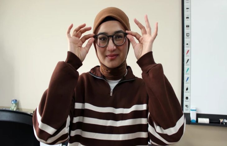 Genç bir kadın, kahverengi tonlarda çizgili bir üst ve başörtüsüyle sınıf benzeri bir ortamda duruyor. Yuvarlak çerçeveli gözlüğünü iki eliyle düzeltiyor ve hafifçe gülümsüyor. Arka planda bir beyaz tahta ve eğitim materyalleri görülüyor.