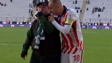 Stadyumda, bir futbolcu forma ve kaptan bandıyla sahada dururken, yanında bulunan taraftarın kulağına eğilip samimi bir şekilde konuşuyor. Taraftar atkı ve şapka takmış; arka planda tribünler ve saha atmosferi görülüyor.