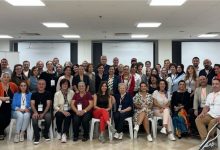 Geniş bir salonda, farklı yaş gruplarından kalabalık bir ekip toplu fotoğraf için bir araya gelmiş. Ön sırada sandalyede oturanlar, arkada ayakta duranlarla birlikte kameraya gülümserken çoğunun boynunda etkinlik kartları bulunuyor. Ortam, bir zirve ya da toplantı sonrası çekilmiş hatıra fotoğrafı havası taşıyor.