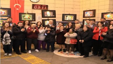 Bir iç mekânda, duvarda Türk bayrağı ve saat bulunan alanda kalabalık bir grup yan yana dizilmiş halde alkış tutuyor. Önlerinde farklı yaşlardan bireyler yer alırken, arkada çerçevelenmiş renkli resimler sergileniyor. Ortam, bir sergi ya da etkinlik açılışını andırıyor.