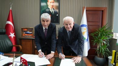 Bir makam odasında çekilmiş bir kare. İki erkek takım elbiseyle bir masanın üzerinde yer alan belgeleri imzalarken öne doğru eğilmiş poz veriyor. Masada evraklar, kalemler ve küçük bir Türk bayrağı bulunuyor. Arkada duvarda Mustafa Kemal Atatürk’ün portresi asılı. Odanın bir yanında Türk bayrağı, diğer yanında kurumsal bir bayrak yer alıyor. Resmî bir protokol imza töreni atmosferi hâkim.