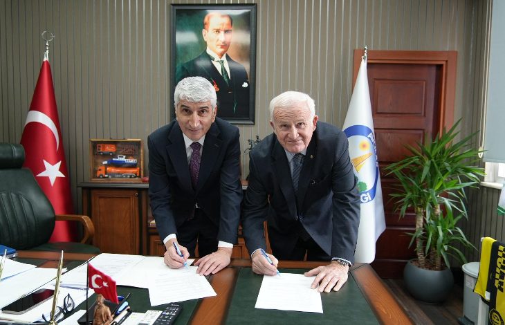 Bir makam odasında çekilmiş bir kare. İki erkek takım elbiseyle bir masanın üzerinde yer alan belgeleri imzalarken öne doğru eğilmiş poz veriyor. Masada evraklar, kalemler ve küçük bir Türk bayrağı bulunuyor. Arkada duvarda Mustafa Kemal Atatürk’ün portresi asılı. Odanın bir yanında Türk bayrağı, diğer yanında kurumsal bir bayrak yer alıyor. Resmî bir protokol imza töreni atmosferi hâkim.