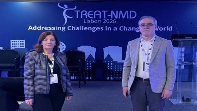 Görselde, bir konferans salonunda sahne önünde duran iki kişi görülüyor. Arkadaki büyük ekranda “TREAT-NMD Lisbon 2026 – Addressing Challenges in a Changing World” yazısı yer alıyor. Kadın ve erkek katılımcılar yaka kartlarıyla kamera karşısında poz verirken, sahnede panel koltukları ve konuşma kürsüsü de dikkat çekiyor. Bu kare, uluslararası bir konferans etkinliği sırasında çekilmiş gibi görünüyor.