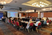 Bir otel toplantı salonunda düzenlenen atölye çalışması görülüyor. U düzeninde yerleştirilmiş masalar etrafında çoğunluğu kadınlardan oluşan katılımcılar oturuyor, not alıyor ve sunumu dinliyor. Ön tarafta projeksiyon ekranında “Engelli Kadın Derneği” sunumu yer alırken, yanında Avrupa Birliği bayrağı ve proje afişleri bulunuyor. Ortam resmi ama aynı zamanda etkileşimli; tartışma ve öğrenme havası hâkim.