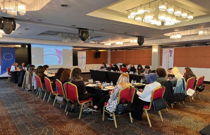 Bir otel toplantı salonunda düzenlenen atölye çalışması görülüyor. U düzeninde yerleştirilmiş masalar etrafında çoğunluğu kadınlardan oluşan katılımcılar oturuyor, not alıyor ve sunumu dinliyor. Ön tarafta projeksiyon ekranında “Engelli Kadın Derneği” sunumu yer alırken, yanında Avrupa Birliği bayrağı ve proje afişleri bulunuyor. Ortam resmi ama aynı zamanda etkileşimli; tartışma ve öğrenme havası hâkim.