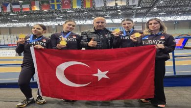 Kapalı bir spor salonunda çekilmiş fotoğrafta, “Türkiye” yazılı spor kıyafetleri giyen dört kadın sporcu ve ortalarında duran bir erkek görülüyor. Sporcular boyunlarında altın madalyalarını gösterirken birlikte büyük bir Türk bayrağı tutuyor. Arka planda pist alanı ve farklı ülkelere ait bayraklar yer alıyor. Görsel, uluslararası bir spor organizasyonunda kazanılan başarıyı yansıtan bir anı gösteriyor.