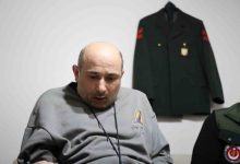 İbrahim Taş, duvarda asılı üniforma önünde oturuyor.