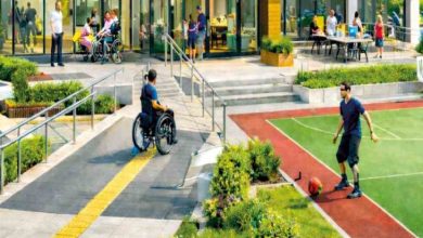 İllüstrasyonlu görselde, modern bir binanın önünde yer alan **engelsiz ve erişilebilir şekilde tasarlanmış bir açık alan** görülmektedir; rampalar ve sarı yönlendirme yolu sayesinde tekerlekli sandalye kullanan bireyler rahatça hareket ederken, bazı insanlar bahçede oturup sohbet etmekte ve bir kişi de yan taraftaki spor alanında top oynamaktadır.