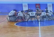 Bir spor salonunda, tekerlekli sandalyede basketbol oynayan sporcular maç öncesi ya da mola anında çember şeklinde toplanmış. Oyuncular omuz omuza vererek takım ruhunu pekiştiriyor. Forma numaraları görünüyor, saha çizgileri net; atmosfer rekabetten çok birlik ve motivasyon hissi taşıyor.