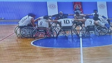 Bir spor salonunda, tekerlekli sandalyede basketbol oynayan sporcular maç öncesi ya da mola anında çember şeklinde toplanmış. Oyuncular omuz omuza vererek takım ruhunu pekiştiriyor. Forma numaraları görünüyor, saha çizgileri net; atmosfer rekabetten çok birlik ve motivasyon hissi taşıyor.