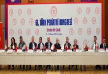 Görselde, 61. Türk Pediatri Kongresi için hazırlanmış bir salonda uzun bir masada yan yana oturan konuşmacılar görülüyor. Masanın üzerinde Türkiye ve kurum bayrakları, isimlikler ve mikrofonlar bulunuyor. Arka planda kongrenin adı, logosu ve “Umudun Adı: Çocuk – 15-19 Nisan 2026, Antalya” yazılı büyük bir pano yer alıyor. Ortam resmi, akademik ve kongre atmosferini yansıtıyor.