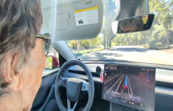 Direksiyon başında yaşlı bir kadın görülüyor. Araç içinde büyük bir dijital ekran var ve yolda ilerlerken sürüş destek sistemi açık görünüyor. Dışarıda ağaçlık bir yol uzanıyor.