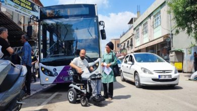 Bir şehir sokağında, belediye otobüsünün önünde tekerlekli sandalye kullanan bir kişi duruyor. Yanında bir kadın elinde kâğıtlarla onunla konuşuyor. Etrafta park halindeki araçlar ve olayı izleyen birkaç kişi var. Sahne, ulaşım sırasında yaşanan bir sorun ve buna verilen tepkiyi yansıtıyor.