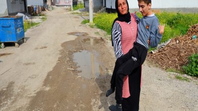 Bozuk ve çamurlu bir mahalle yolunda, bir kadın kucağında bir çocuğu taşıyor. Etrafta su birikintileri ve bakımsızlık dikkat çekerken, arka planda evler ve boş alanlar görülüyor.