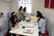 Aydınlık bir atölye ortamında, birkaç kadın uzun bir masa etrafında oturmuş el işi yapıyor. Masanın üzerinde boyalı tepsiler, süsleme malzemeleri ve çeşitli sanat ürünleri bulunuyor. Arka tarafta iki kişi ayakta çalışırken, diğerleri dikkatle detaylara odaklanmış. Ortam sakin, üretken ve paylaşım dolu bir atölye havası taşıyor.