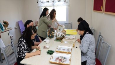 Aydınlık bir atölye ortamında, birkaç kadın uzun bir masa etrafında oturmuş el işi yapıyor. Masanın üzerinde boyalı tepsiler, süsleme malzemeleri ve çeşitli sanat ürünleri bulunuyor. Arka tarafta iki kişi ayakta çalışırken, diğerleri dikkatle detaylara odaklanmış. Ortam sakin, üretken ve paylaşım dolu bir atölye havası taşıyor.