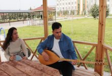 Görselde, bir kamelya içinde oturan iki kişi görülüyor. Erkek kişi bağlama çalarken yanında oturan kadın da def tutuyor. Arka planda yeşil bir bahçe, ağaçlar ve bir bina dikkat çekiyor. İkili, açık havada sakin bir ortamda müzikle ilgileniyor gibi görünüyor.