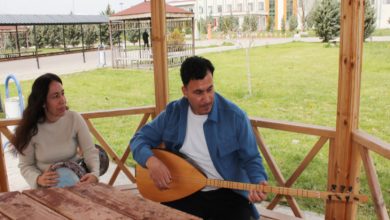 Görselde, bir kamelya içinde oturan iki kişi görülüyor. Erkek kişi bağlama çalarken yanında oturan kadın da def tutuyor. Arka planda yeşil bir bahçe, ağaçlar ve bir bina dikkat çekiyor. İkili, açık havada sakin bir ortamda müzikle ilgileniyor gibi görünüyor.