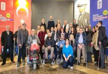 Kalabalık bir grup, kapalı bir salonda toplu fotoğraf için poz veriyor. Aralarında tekerlekli sandalye kullananlar ve bastonla ayakta duranlar var. Arkada Türk bayrağı ve Mustafa Kemal Atatürk’ün portresi yer alıyor. Katılımcılar resmi bir etkinlik sonrası bir araya gelmiş gibi görünüyor.