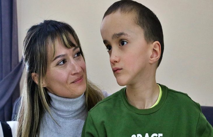 Bir odada çekilmiş fotoğrafta, kısa saçlı küçük bir çocuk yana doğru bakarken, yanında oturan bir kadın ona sevgi ve dikkatle bakıyor. Kadının yüzünde hafif bir tebessüm var; aralarındaki bağ sıcak ve koruyucu bir his veriyor. Çocuk ise düşünceli ve biraz dalgın görünüyor.