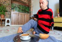 Ev ortamında yerde oturan bir adam, ayaklarını kullanarak bir tepsideki çaya uzanıyor. Üzerinde kırmızı-siyah çizgili bir kazak var. Yanında ekmek bulunuyor; arka planda akvaryum, dolap ve bitkiler yer alıyor. Sahne, günlük yaşamını ayaklarıyla sürdüren birinin güçlü ve bağımsız anını yansıtıyor.