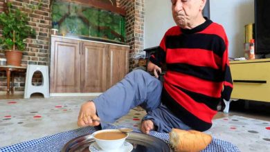 Ev ortamında yerde oturan bir adam, ayaklarını kullanarak bir tepsideki çaya uzanıyor. Üzerinde kırmızı-siyah çizgili bir kazak var. Yanında ekmek bulunuyor; arka planda akvaryum, dolap ve bitkiler yer alıyor. Sahne, günlük yaşamını ayaklarıyla sürdüren birinin güçlü ve bağımsız anını yansıtıyor.