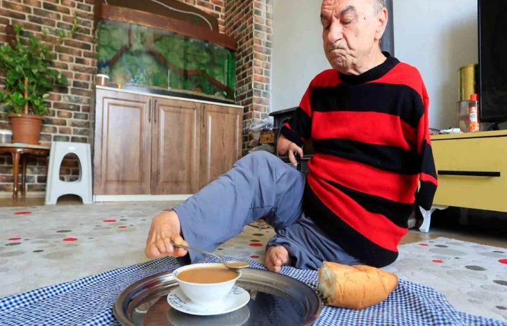 Ev ortamında yerde oturan bir adam, ayaklarını kullanarak bir tepsideki çaya uzanıyor. Üzerinde kırmızı-siyah çizgili bir kazak var. Yanında ekmek bulunuyor; arka planda akvaryum, dolap ve bitkiler yer alıyor. Sahne, günlük yaşamını ayaklarıyla sürdüren birinin güçlü ve bağımsız anını yansıtıyor.