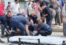 Bir sokakta yaşanan kazaya müdahale anı görülüyor. Sağlık ekipleri yerde yatan bir kişiye ilk yardım yaparken, çevrede toplanan kalabalık endişeli şekilde izliyor. Ambulans sedyesi hazır bekletiliyor; ortam gergin ve telaşlı.