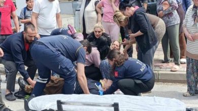 Bir sokakta yaşanan kazaya müdahale anı görülüyor. Sağlık ekipleri yerde yatan bir kişiye ilk yardım yaparken, çevrede toplanan kalabalık endişeli şekilde izliyor. Ambulans sedyesi hazır bekletiliyor; ortam gergin ve telaşlı.