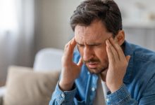 Bir adam, ellerini şakaklarına götürmüş halde baş ağrısı ya da baş dönmesi yaşıyor gibi görünüyor. Ev ortamında çekilen kare, yorgunluk, stres veya sağlık sorunlarının günlük hayattaki etkisini güçlü biçimde yansıtıyor.