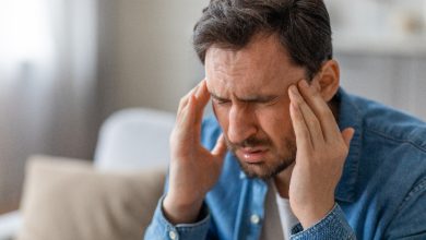 Bir adam, ellerini şakaklarına götürmüş halde baş ağrısı ya da baş dönmesi yaşıyor gibi görünüyor. Ev ortamında çekilen kare, yorgunluk, stres veya sağlık sorunlarının günlük hayattaki etkisini güçlü biçimde yansıtıyor.