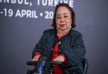 Tekerlekli sandalye kullanan bir kadın, bir etkinlik alanında kameraya dönük şekilde poz veriyor. Arkada İngilizce yazılar ve tarih bilgisi yer alıyor; ortam bir konferans ya da uluslararası toplantıyı çağrıştırıyor. Kadının sakin ama güçlü bir ifadesi var, resmi bir katılımcı gibi duruyor.