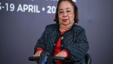 Tekerlekli sandalye kullanan bir kadın, bir etkinlik alanında kameraya dönük şekilde poz veriyor. Arkada İngilizce yazılar ve tarih bilgisi yer alıyor; ortam bir konferans ya da uluslararası toplantıyı çağrıştırıyor. Kadının sakin ama güçlü bir ifadesi var, resmi bir katılımcı gibi duruyor.