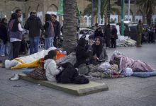Açık alanda, yerde serili yatak ve battaniyeler üzerinde oturan bir aile görülüyor. Kadınlar ve çocuklar sokakta kalmış; etrafta eşyalar dağınık. Arka planda kalabalık insanlar ve hareketli bir şehir ortamı dikkat çekiyor.