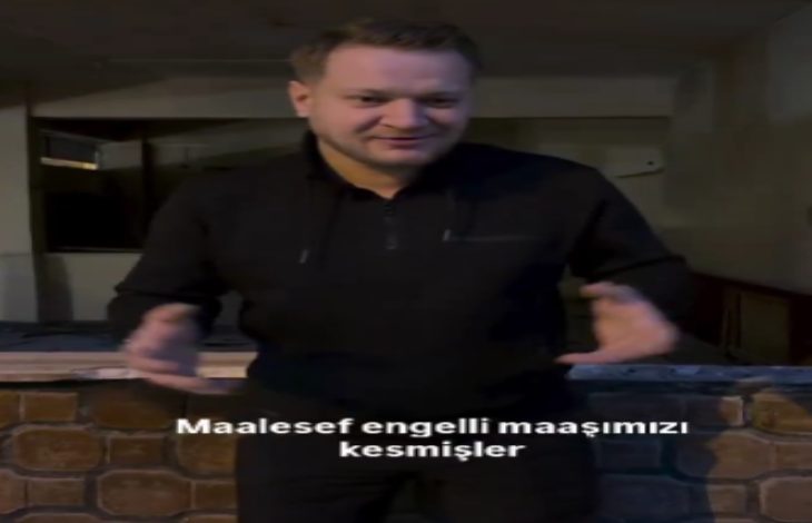 Kapalı bir mekânda ayakta duran bir adam, ellerini kullanarak bir şeyi anlatır gibi konuşuyor. Üzerinde siyah kıyafetler var. Görüntünün alt kısmında “Maalesef engelli maaşımızı kesmişler” yazısı yer alıyor. Sahne, yaşadığı bir mağduriyeti dile getirdiği bir anı yansıtıyor.