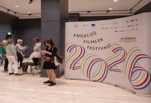 Bir etkinlik alanında, büyük bir pano üzerinde “Engelsiz Filmler Festivali 2026” yazısı yer alıyor. Renkli grafiklerle tasarlanan afişin önünde birkaç kişi ayakta sohbet ediyor ya da telefonlarına bakıyor. Ortam modern ve sakin; festival havası hissediliyor.