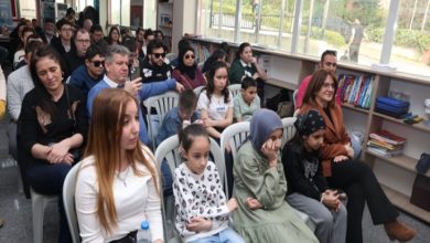 Bir salon ortamında, farklı yaş gruplarından insanların sandalyelerde yan yana oturarak bir etkinliği izlediği görülüyor. Ön sıralarda çocuklar, arka sıralarda yetişkinler yer alıyor. Yan tarafta kitaplık ve geniş pencereler bulunuyor. Sahne, toplu bir etkinlik ya da dinleti sırasında izleyicilerin dikkatle odaklandığı bir anı yansıtıyor.