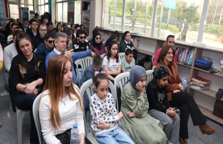 Bir salon ortamında, farklı yaş gruplarından insanların sandalyelerde yan yana oturarak bir etkinliği izlediği görülüyor. Ön sıralarda çocuklar, arka sıralarda yetişkinler yer alıyor. Yan tarafta kitaplık ve geniş pencereler bulunuyor. Sahne, toplu bir etkinlik ya da dinleti sırasında izleyicilerin dikkatle odaklandığı bir anı yansıtıyor.